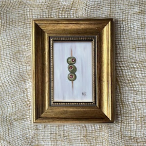 May include: A small framed painting featuring three green olives with red pimentos on a wooden skewer against a white background. The artwork is enclosed in an ornate, metallic gold frame with a textured border and includes the initials MK in the corner.