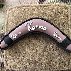 May include: A pink boomerang with black edges and the text "Karma Returns" on both sides. The boomerang is on a brown fabric background.