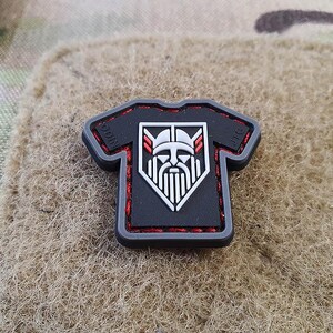 May include: Black and red embroidered patch with a white graphic of a viking wearing a helmet and holding an axe. The patch is shaped like a t-shirt and has the text "ODIN" and "JIG" on the shoulders.
