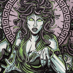 Puede incluir: Parche bordado con una ilustración verde y negra de Medusa, una figura mitológica griega con serpientes por cabello, rodeada de un patrón de clave griega morado y blanco.