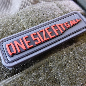 May include: A grey patch with the text "ONE SIZE FITS ALL" in black and red lettering. The patch has a hook and loop backing.