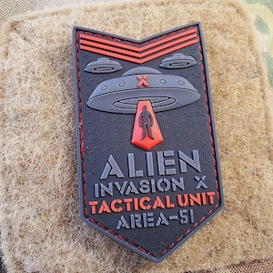 May include: A black and red embroidered patch with a UFO, a stick figure, and the text "Alien Invasion X Tactical Unit Area-51".