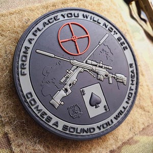 Puede incluir: Un parche negro y gris con un contorno blanco. El parche presenta una imagen estilizada de dos rifles cruzados con una carta de juego con un símbolo de pica. El texto en el parche dice "From a place you will not see comes a sound you will not hear".