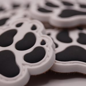 Könnte beinhalten: Nahaufnahme von mehreren weißen, pfotenabdruckförmigen Objekten mit schwarzen Ballen. Die Pfotenabdrücke sind gruppiert, einige sind scharf, andere unscharf. Die Objekte scheinen aus einem glatten Material zu bestehen.