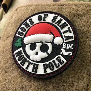 Op de afbeelding: Een zwart-witte patch met de tekst "Sons of Santa North Pole" en een schedel die een rood-witte kerstmanmuts draagt. De patch heeft een rode rand en witte stiksels.