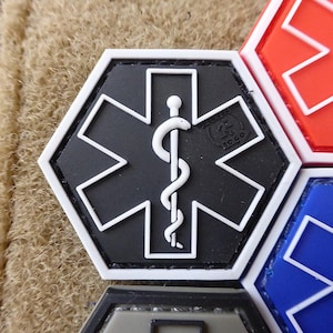 Peut inclure: Un patch médical noir et blanc avec une étoile de vie et un serpent enroulé autour d'un bâton. Le patch est hexagonal et a une bordure blanche. Le texte "Tactical TO GO" est imprimé sur le patch.