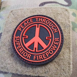 Pode incluir: Um remendo preto e vermelho com um sinal de paz no centro. O texto ao redor do sinal de paz diz "Peace Through Superior Firepower".