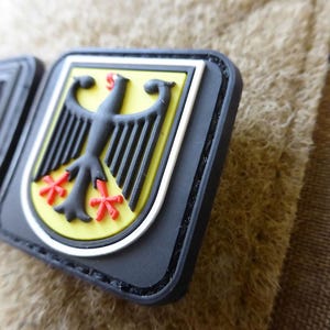 May include: A black, yellow, and red emblem featuring a stylized eagle, the national symbol of Germany. The emblem is set against a black background with a white border. The patch is attached to a camouflage fabric.