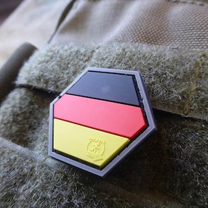 Puede incluir: Un parche hexagonal negro, rojo y amarillo con la bandera alemana y el texto "Made in Germany" sobre un fondo de tela marrón.
