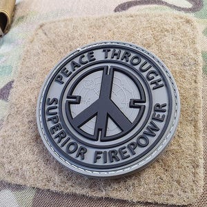 Peut inclure: Un patch gris et noir avec un signe de paix et le texte "Peace Through Superior Firepower".