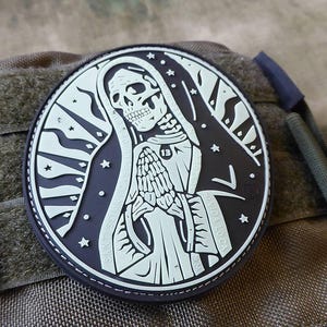 Könnte beinhalten: Runder, schwarz-weißer Aufnäher mit einem Skelett in einer Kapuzenrobe, die Hände zum Gebet gefaltet. Das Design umfasst Sterne und Text. Die Zahl "13" ist sichtbar. Der Aufnäher befindet sich vor einem strukturierten, olivgrünen Hintergrund.