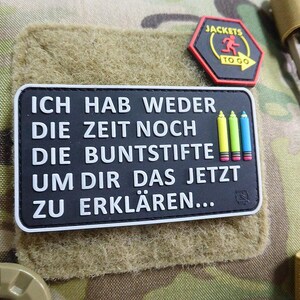 May include: A black rectangular patch with white text that reads "ICH HAB WEDER DIE ZEIT NOCH DIE BUNTSTIFTE UM DIR DAS JETZT ZU ERKLÄREN...". The patch has three colored pencil graphics. A red, black, and yellow patch is above it.