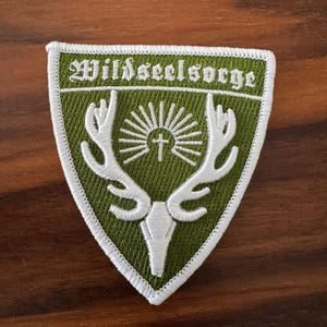 May include: Embroidered patch in a shield shape with the text "Wildseelsorge" in green. The patch features a white deer head with antlers, a cross, and sun rays on a green background, outlined in white.