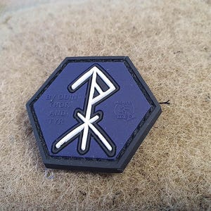 Könnte beinhalten: Ein blauer und weißer sechseckiger Patch mit einem weißen Symbol und dem Text "By Odin, Thor and Tyr" und "Jackets To Go" in weiß.