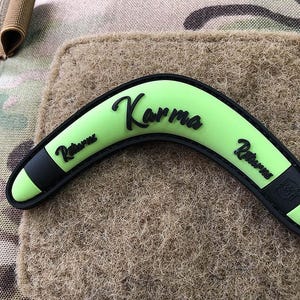 Può includere: Un boomerang verde e nero con la parola "Karma" scritta su di esso. Il boomerang è fatto di un materiale morbido e flessibile e ha un bordo nero attorno ai bordi.