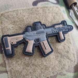 Könnte beinhalten: Ein schwarzer und brauner Patch, der ein stilisiertes Bild eines Gewehrs mit dem Text "STEU" auf der Seite zeigt.