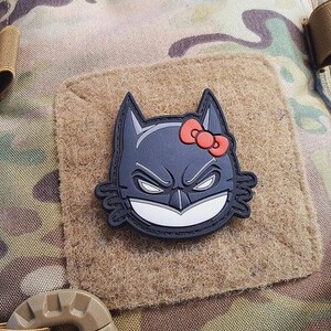 Puede incluir: Un parche negro y gris con un diseño híbrido de Batman y Hello Kitty. El parche tiene un lazo rojo y está fijado a un cuadrado de tela beige, sobre un fondo de camuflaje.