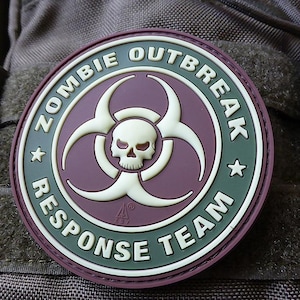 Puede incluir: Un parche circular con el texto "ZOMBIE OUTBREAK RESPONSE TEAM" en letras blancas. El parche presenta una calavera y un símbolo de peligro biológico en el centro, con una combinación de colores púrpura y verde.