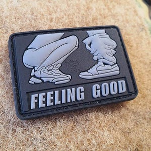 Op de afbeelding: Een rechthoekige, zwarte patch met een grijze afbeelding van benen en sneakers. De tekst "FEELING GOOD" staat in grijze, vette letters onderaan. De patch ligt op een getextureerd, bruin oppervlak.