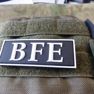 May include: A black and white patch with the letters "BFE" in white on a black background. The patch is attached to a green tactical bag.