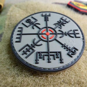 Könnte beinhalten: Runder, grauer Aufnäher mit einem schwarzen Vegvisir-Symbol und einem roten Zentrum. Der Aufnäher hat einen schwarzen Rand und befindet sich auf einem Tarnhintergrund. Das Vegvisir ist ein Symbol der Führung.