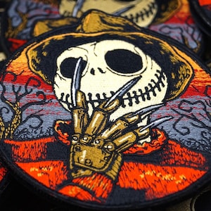 Könnte beinhalten: Ein schwarzer und orangefarbener gestickter Aufnäher mit einer Figur mit einem zusammengenähten Gesicht und einem Freddy-Krueger-artigen Handschuh. Die Figur trägt einen braunen Hut und ist vor einem rot-orange-grauen Hintergrund platziert.