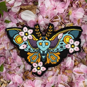 Könnte beinhalten: Ein farbenfroher gestickter Aufnäher mit einer Motte mit floralen Akzenten. Die Motte hat gelbe, orange, blaue und violette Flügel mit schwarzen Umrissen. Die Blumen sind rosa und weiß mit gelben Zentren. Der Aufnäher befindet sich auf einem Hintergrund aus rosa Blütenblättern.