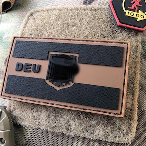 Könnte beinhalten: Ein braunes und schwarzes Patch mit dem Wort "DEU" und einem schwarzen Adler-Design. Das Patch ist auf ein Tarnstoff genäht.