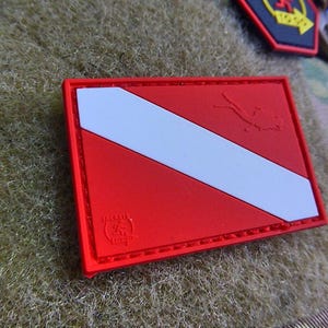 Könnte beinhalten: Roter und weißer Taucherflaggen-Patch mit einer genähten Umrandung. Der Patch hat einen weißen diagonalen Streifen auf rotem Hintergrund. Der Patch hat ein kleines Logo in der linken unteren Ecke.