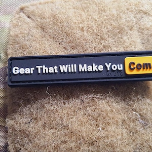 Può includere: Una toppa rettangolare nera con testo bianco che recita "Gear That Will Make You Come" e un rettangolo giallo con la parola "Come". La toppa è su una superficie strutturata color beige.