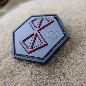 Może przedstawiać: Szesciokątna naszywka w kolorze czarnym i szarym z czerwonym stylizowanym symbolem w środku. Symbol to stylizowane "X" z linią przechodzącą przez środek. Tekst "JACKETS TOGO" jest nadrukowany na szaro pod symbolem.