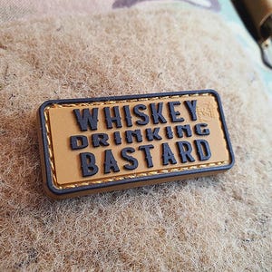Può includere: Un cerotto marrone con cuciture nere e il testo "Whiskey Drinking Bastard" in lettere nere.