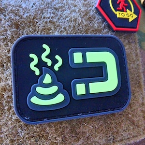 May include: A black patch with a green poop emoji and a green magnet. The poop emoji has steam coming out of it.