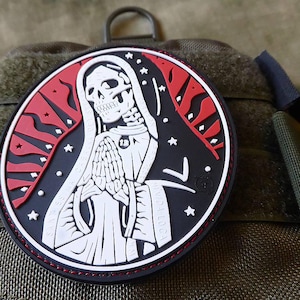 May include: A black and red patch featuring a white image of a skeleton wearing a hooded robe. The skeleton is holding its hands together in a prayer position. The patch has a red and white flame design around the edges.
