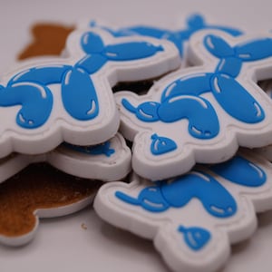 Könnte beinhalten: Ein Haufen weiß-blauer Ballonhund-Aufnäher. Die Aufnäher haben einen weißen Umriss und ein blaues Ballonhund-Design. Die Aufnäher sind übereinander gestapelt, wobei etwas braunes, strukturiertes Trägermaterial sichtbar ist.