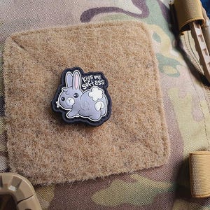 Puede incluir: Un parche de conejo de dibujos animados gris y blanco con el texto "Kiss my soft ass" sobre un cuadrado beige. El parche está sobre un fondo de camuflaje.