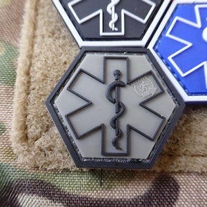 May include: A grey hexagonal patch with a black star of life and a black caduceus symbol. The patch has a black border and a small white circle with the text "ZULU 25 TOGO" in black.