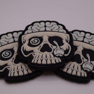 Könnte beinhalten: Drei schwarze und cremefarbene Totenkopf-Patches mit freiliegendem Gehirn. Die Schädel haben ein detailliertes Design mit schwarzem Rand. Augenhöhlen und Zähne sind sichtbar. Die Patches überlappen sich leicht.