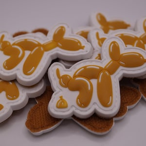 Könnte beinhalten: Eine Ansammlung weißer und gelber Aufnäher in Hundeform. Die Aufnäher zeigen ein Cartoon-Design eines Hundes aus Luftballons. Die Aufnäher liegen auf einem braunen, strukturierten Hintergrund.