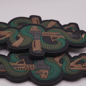 Pode incluir: Vários patches táticos com um design de cobra enrolada em torno de um rifle. Os patches têm uma borda preta, com detalhes verdes, castanhos e cinzentos. A cabeça da cobra é visível e o rifle é detalhado.