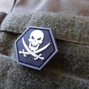 May include: A black and white skull and crossbones patch with a hexagonal shape. The patch has a white skull with crossed swords and a small logo in the center. The patch is attached to a brown and green fabric.