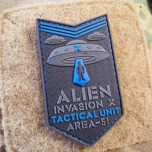 Peut inclure: Patch tactique d'invasion extraterrestre noir et bleu avec un OVNI et un bonhomme en costume bleu. Le texte indique "Alien Invasion X Tactical Unit Area-51".