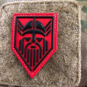 Pode incluir: Patch bordado vermelho e preto com o rosto de um guerreiro viking em forma de escudo.