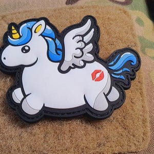 Könnte beinhalten: Ein weißer Einhorn-Aufnäher mit blauen Haaren, einem gelben Horn und grauen Flügeln. Das Einhorn hat einen roten Kussmund auf der Seite und einen schwarzen Umriss. Der Aufnäher befindet sich auf einem beigen Hintergrund.