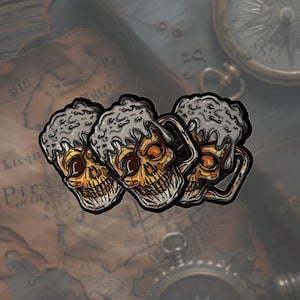 Könnte beinhalten: Drei schädelförmige Bierkrüge mit einem mattierten Glasdesign. Die Krüge sind aus dunkelgrauem Material gefertigt und haben ein goldfarbenes Schädelmotiv.