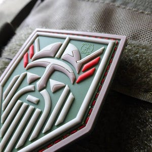 May include: A shield-shaped patch with a stylized face design, including a beard and helmet. The design uses white, red, and grey on a green base, with a brown border. The patch is attached to a textured, olive-green surface.