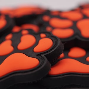 Könnte beinhalten: Nahaufnahme mehrerer schwarzer und orangefarbener Pfotenabdruck-Patches. Die Patches haben einen schwarzen Rand mit orangefarbenen Pfotenballen. Die Patches sind wahrscheinlich aus Gummi oder einem ähnlichen Material.