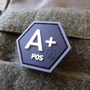 Könnte beinhalten: Ein schwarzer sechseckiger Patch mit weißem Text, der "A+" und "POS" lautet. Der Patch hat ein kleines Logo mit der Aufschrift "Jackets To Go" in weißer Schrift.