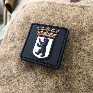 Könnte beinhalten: Ein schwarzer quadratischer Aufnäher mit dem Berliner Wappen. Es zeigt einen schwarzen Bären mit roten Krallen und Zunge auf einem weißen Schild, gekrönt von einer braunen Krone. Der Aufnäher befindet sich auf einem beigefarbenen, strukturierten Hintergrund.