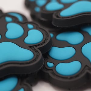 Könnte beinhalten: Nahaufnahme mehrerer schwarzer Pfoten-förmiger Aufnäher mit hellblauen Pfotenabdrücken. Die Aufnäher bestehen aus einem gummiartigen Material und sind übereinander gestapelt. Die Aufnäher sind wahrscheinlich für Tierbesitzer.